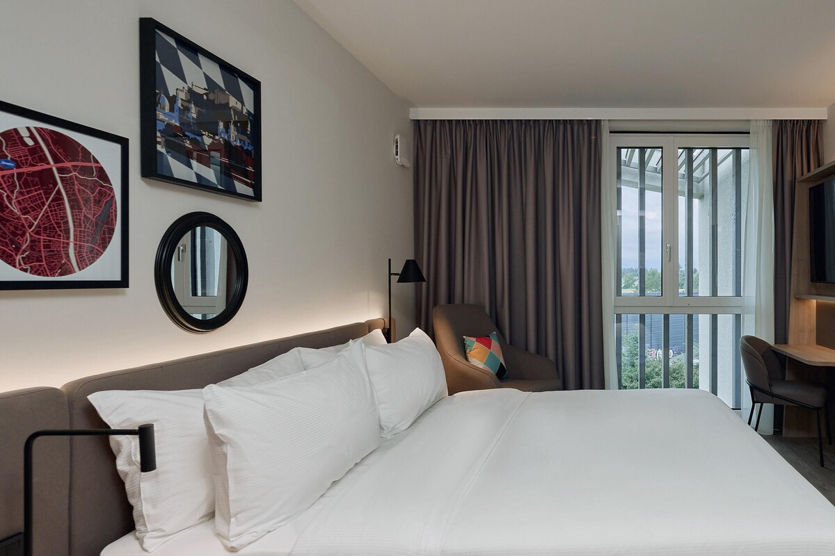 Ein Hotelzimmer mit einem großen Bett, einem Schreibtisch, einem Sessel, einem Fernseher und einem Fenster mit Vorhängen.
