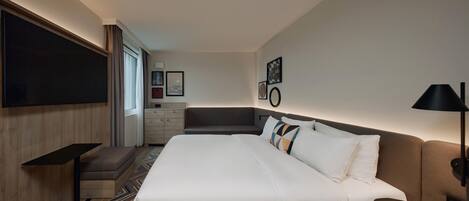 A modern hotel room with a large bed, a desk, a chair, and a TV mounted on the wall.
