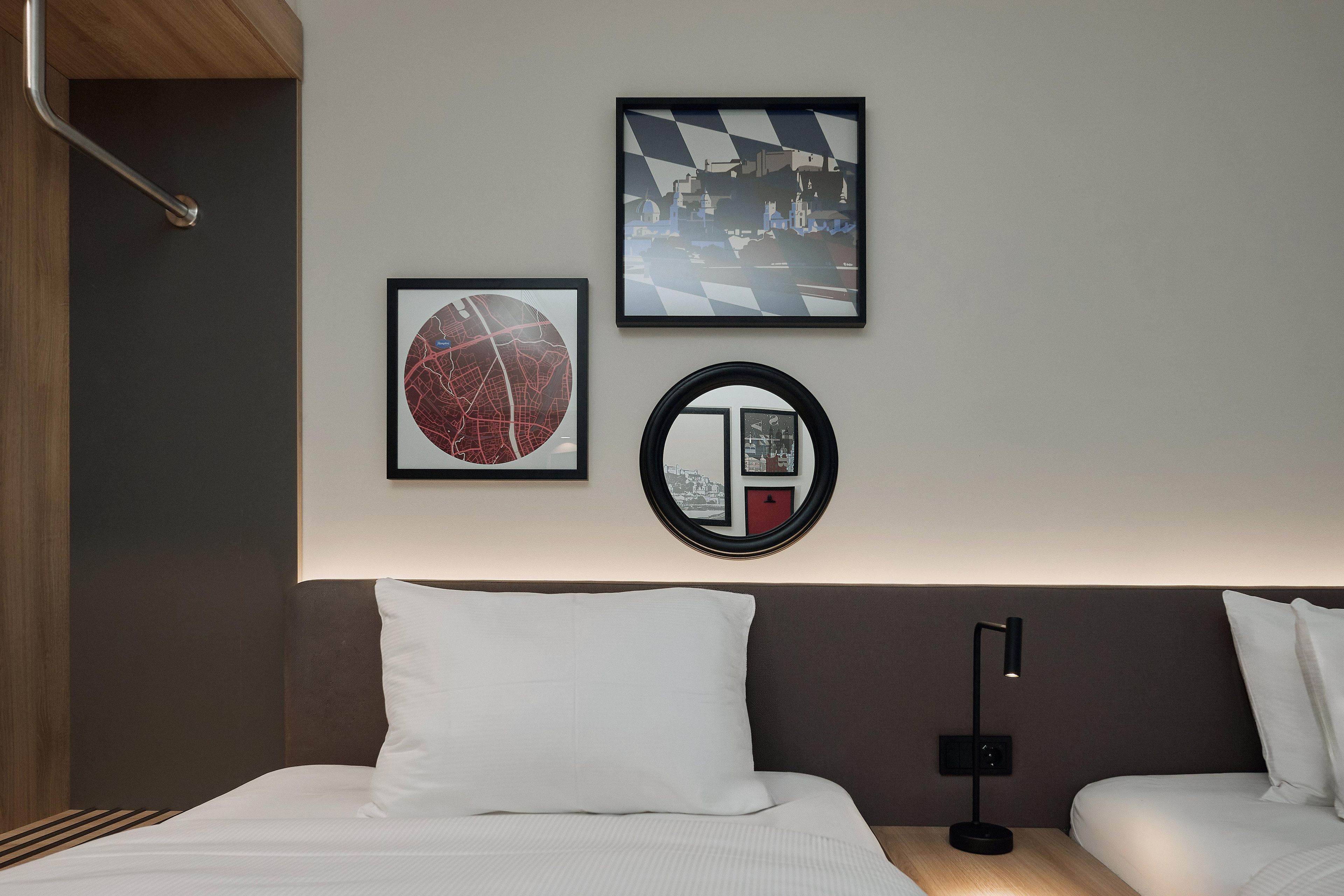A hotel room with a bed, two pillows, a nightstand with a lamp, and three framed artworks on the wall.
