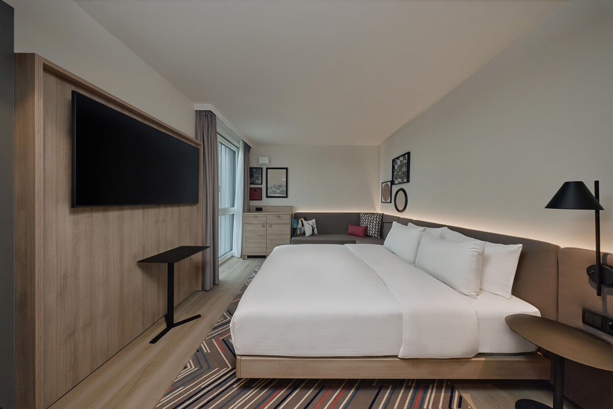 Ein Hotelzimmer mit einem großen Bett, einem an der Wand montierten Fernseher, einem Schreibtisch, einem Sessel und einem Nachttisch.