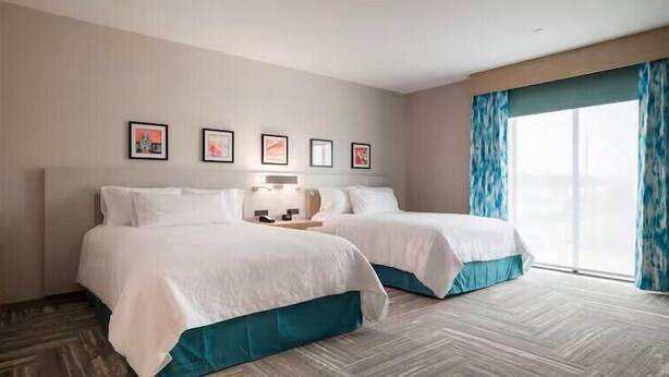 Een hotelkamer met twee bedden, een nachtkastje, een raam met gordijnen en lijstjes met foto's aan de muur.