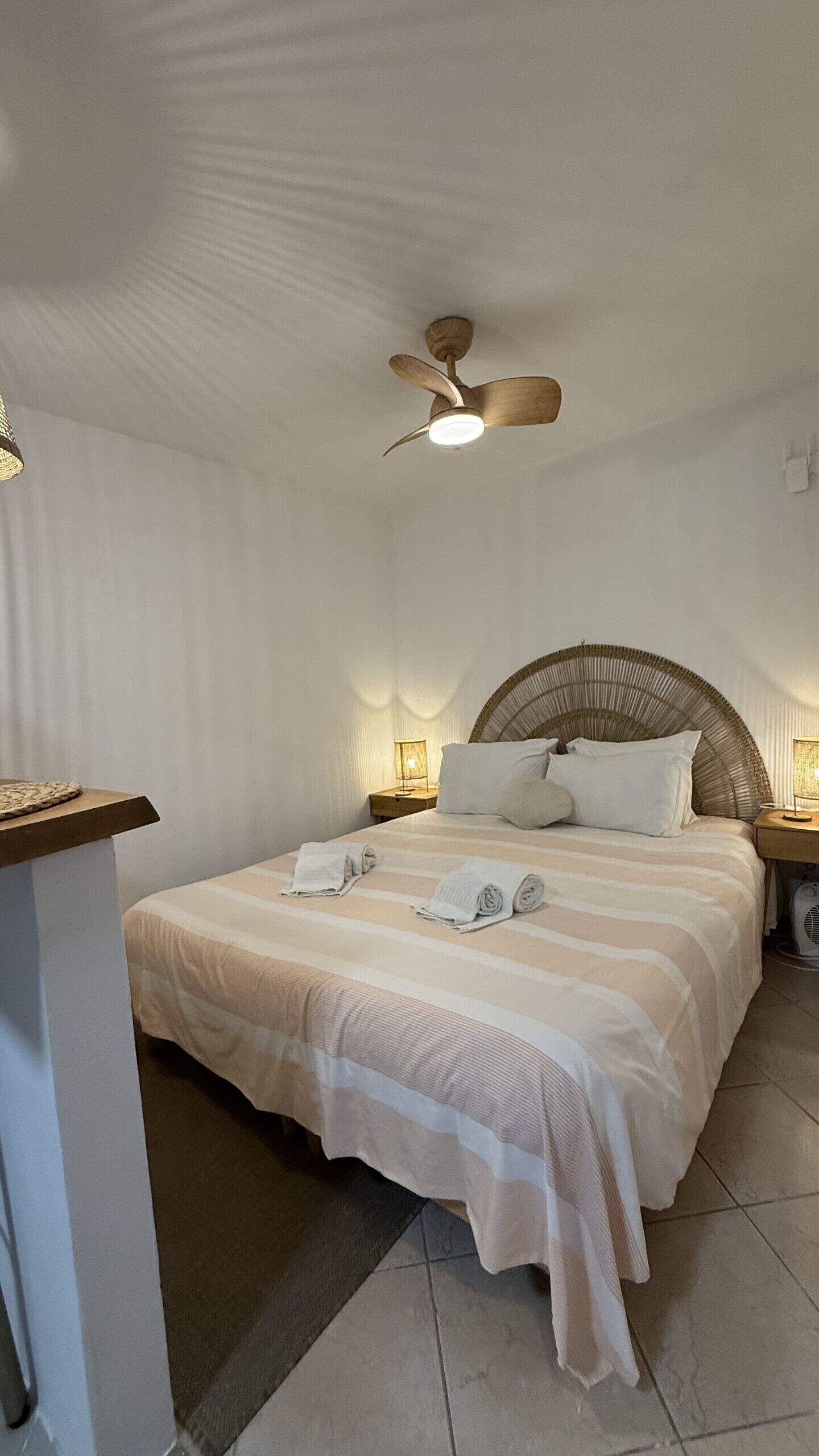 Un dormitorio con cama, ventilador de techo, lámparas de noche y una mesita.