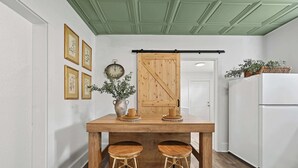 A kitchen with a wooden table, two stools, a hanging wooden door, a white refrigerator, and framed botanical prints on the wall. - The 408 Family Farmhouse (Siloam Springs)