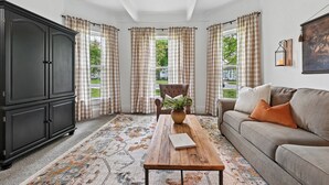 A room with a large dark wooden cabinet, a wooden coffee table with a book, a sofa with cushions, and checkered curtains. - The 408 Family Farmhouse (Siloam Springs)
