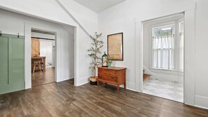 A hallway with wooden flooring, a wooden bench, a small table with a plant, a framed picture, and a window with curtains. - The 408 Family Farmhouse (Siloam Springs)