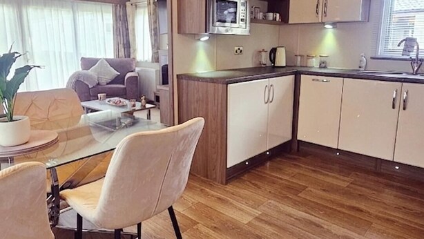 A modern kitchen with wooden cabinets, a built-in oven, and a dining area with a glass table and chairs. - Modern & Spacious 40ft Caravan Sleeps 6, Luxury! (Great Yarmouth)
