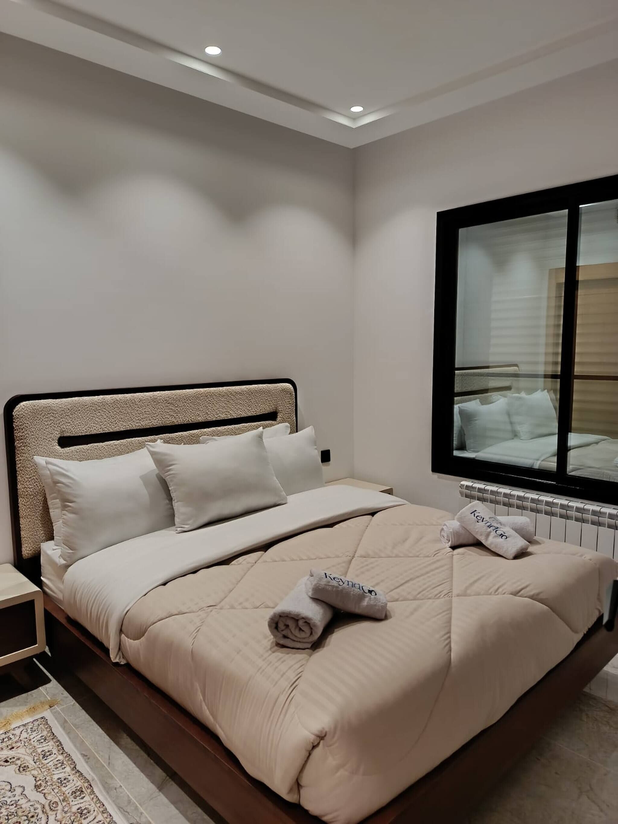 A neatly made bed with a headboard, a window with blinds, and a radiator beneath it.
