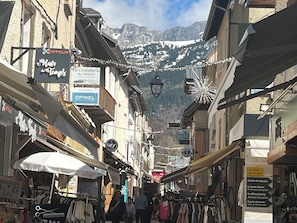 Una calle estrecha con tiendas y letreros, incluyendo 'Hair Tempest' y 'OPINEL', con montañas al fondo.