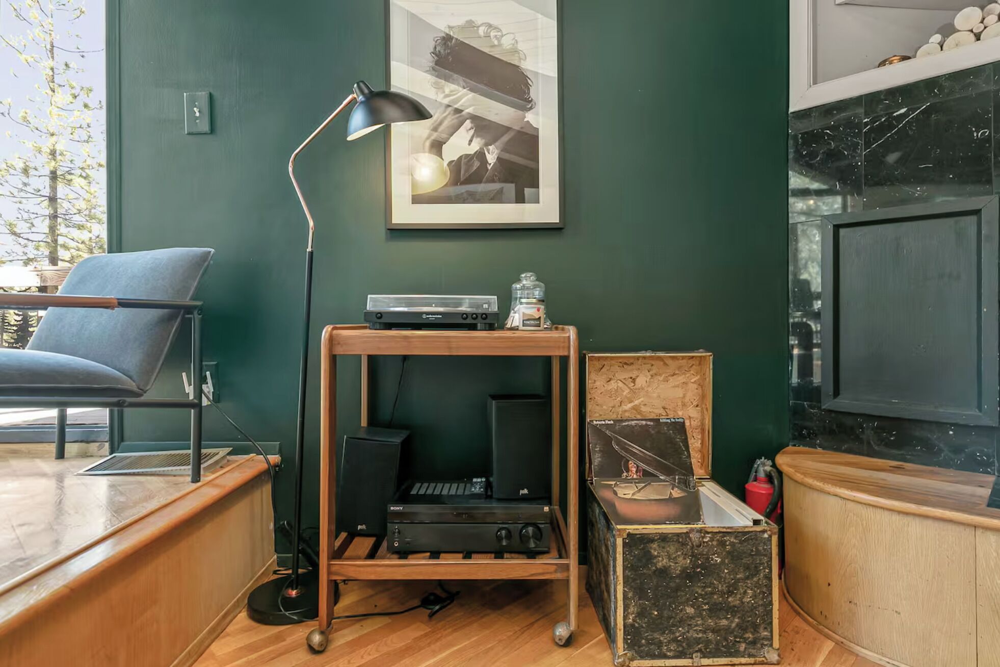 Una habitación con un escritorio de madera, una silla azul, una lámpara de pie negra, un cuadro en la pared y un televisor sobre un mueble.