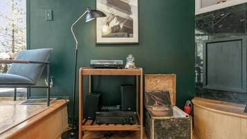 Una habitación con un escritorio de madera, una silla azul, una lámpara de pie negra, un cuadro en la pared y un televisor sobre un mueble.