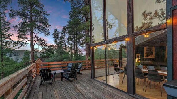 Une terrasse en bois avec des sièges extérieurs et un espace repas fermé par des baies vitrées, offrant une vue sur les arbres et le coucher de soleil.