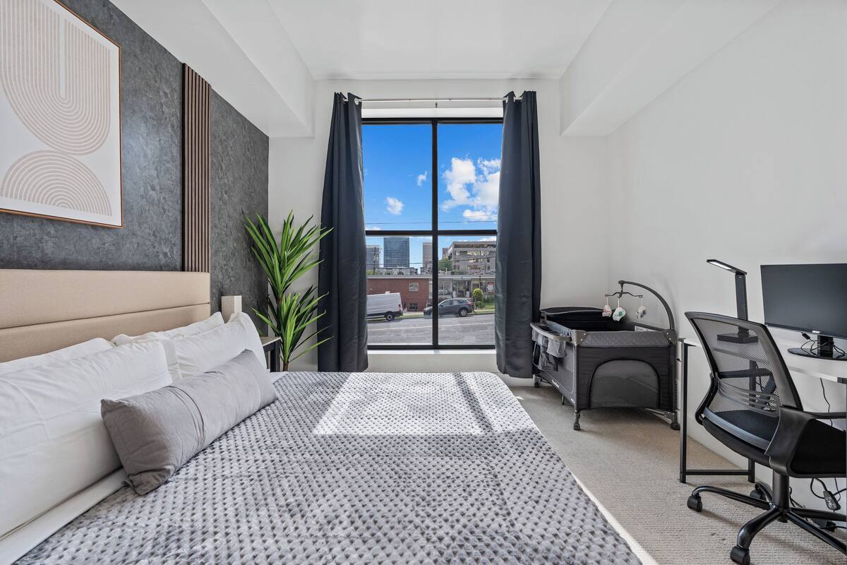 Une chambre d’hôtel moderne, dotée d’un grand lit, d’un bureau avec une chaise, d’une plante en pot et offrant une vue sur la ville grâce à une grande fenêtre.
