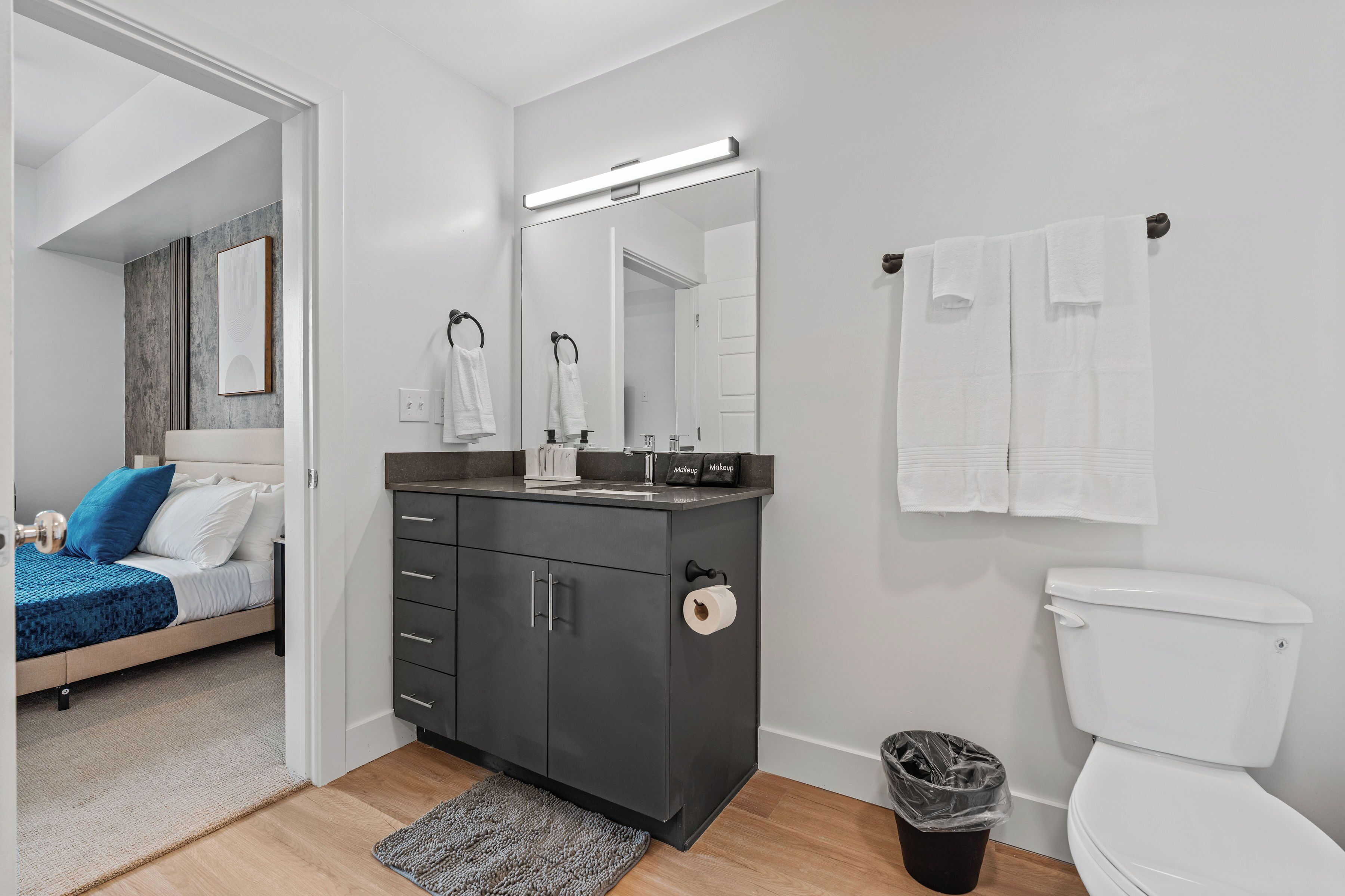 Egy modern fürdőszoba, amelyben WC, mosdókagyló és tükör található. A mellette lévő szobában egy ágy van, kék takaróval és párnákkal.