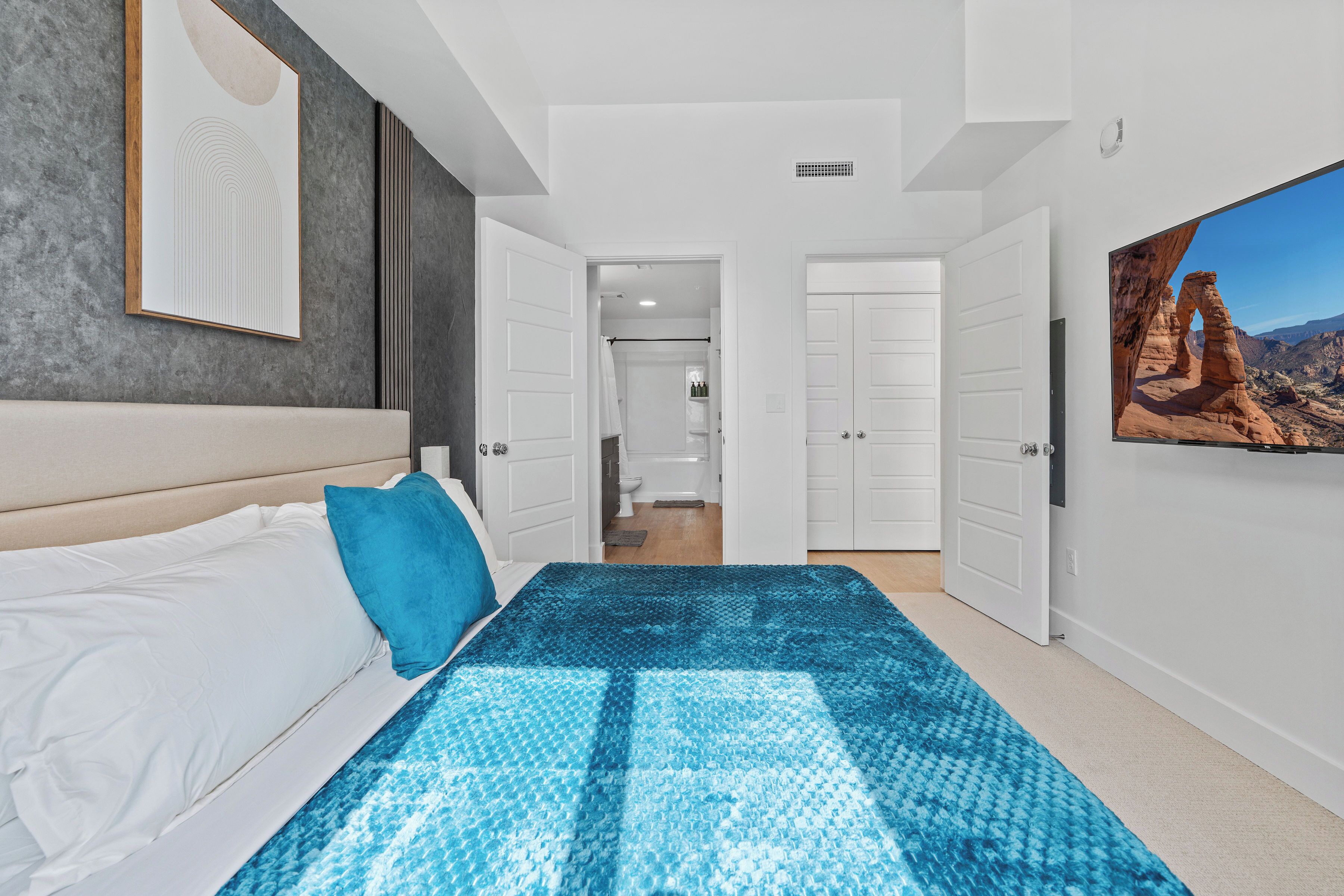 Un dormitorio con cama, una colcha azul estampada, cabecera blanca, almohada azul, una obra de arte enmarcada en la pared y una puerta que da a un baño.