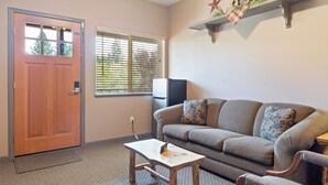 A living room with a wooden door, a window with blinds, a grey sofa with patterned cushions, a small wooden table, and a wall-mounted shelf with decorative items. - SouthFork Lodge (Lowman)