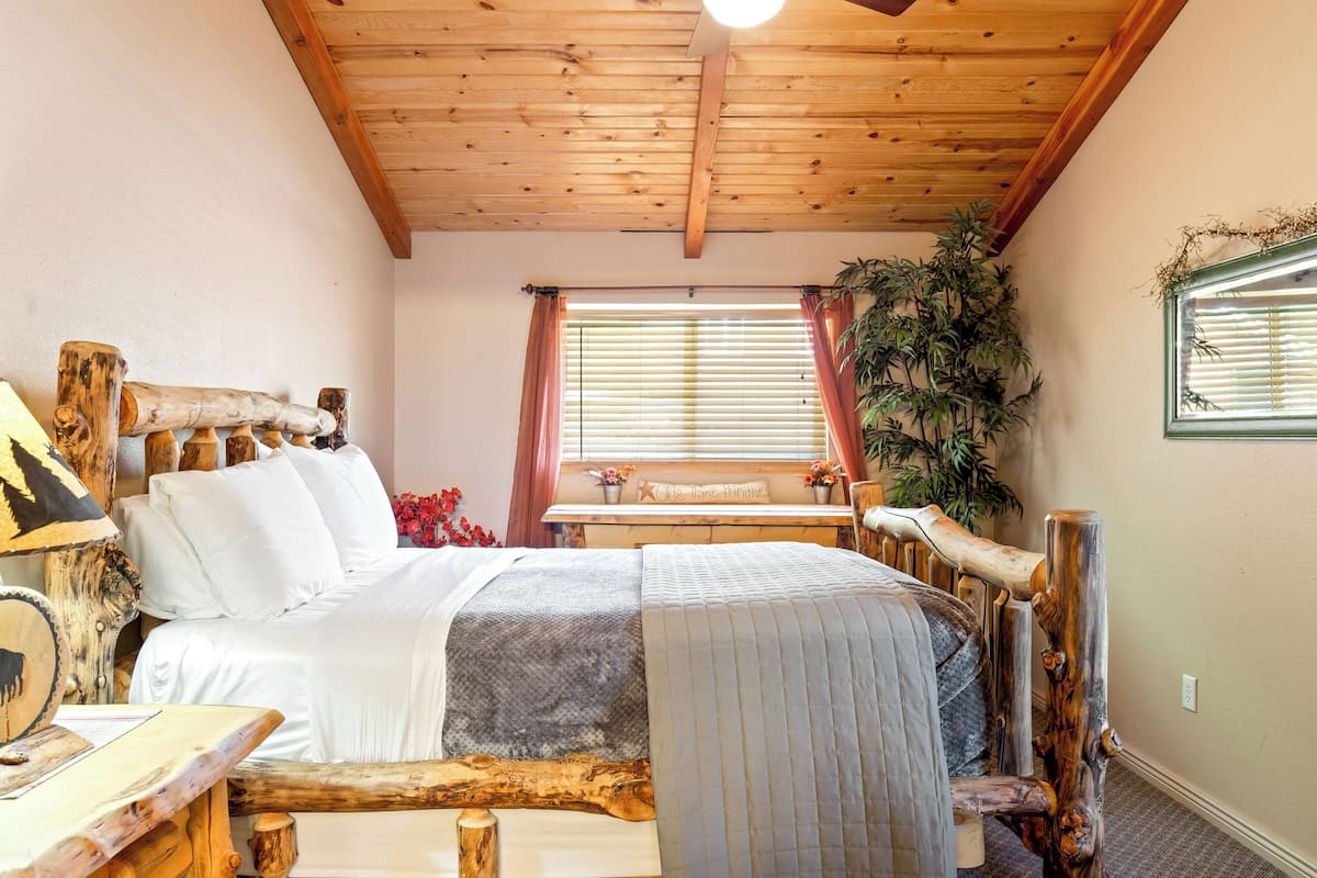 A wooden bed with a log headboard, a bedside table with a lamp, and a window with blinds.