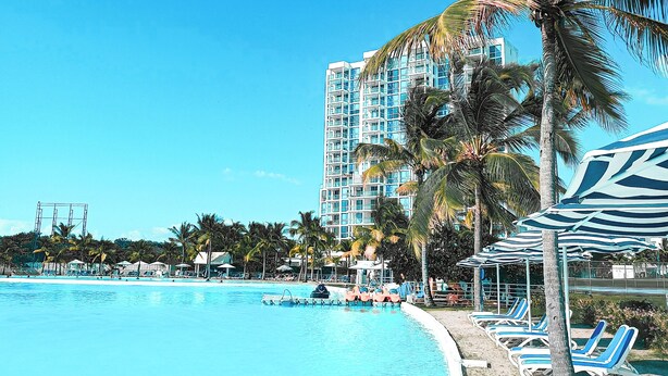A clear blue pool with lounge chairs and palm trees, surrounded by a high-rise building. - Blue Escape Suite (rio hato)