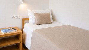 Un lit bien fait, recouvert d’une couverture beige, une table de chevet en bois avec un livre, et une lampe fixée au mur.