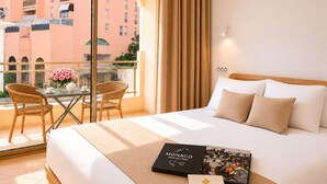 Une chambre d’hôtel avec un lit, une table et des chaises, un balcon donnant sur des immeubles et des rideaux.