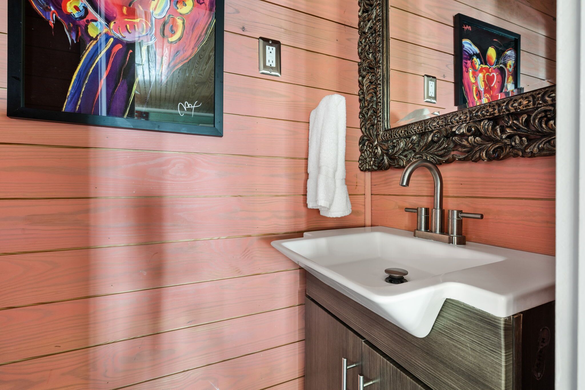 Une salle de bain avec un mur recouvert de panneaux en bois, un lavabo blanc, un miroir doté d’un cadre orné et un tableau placé au-dessus du miroir.