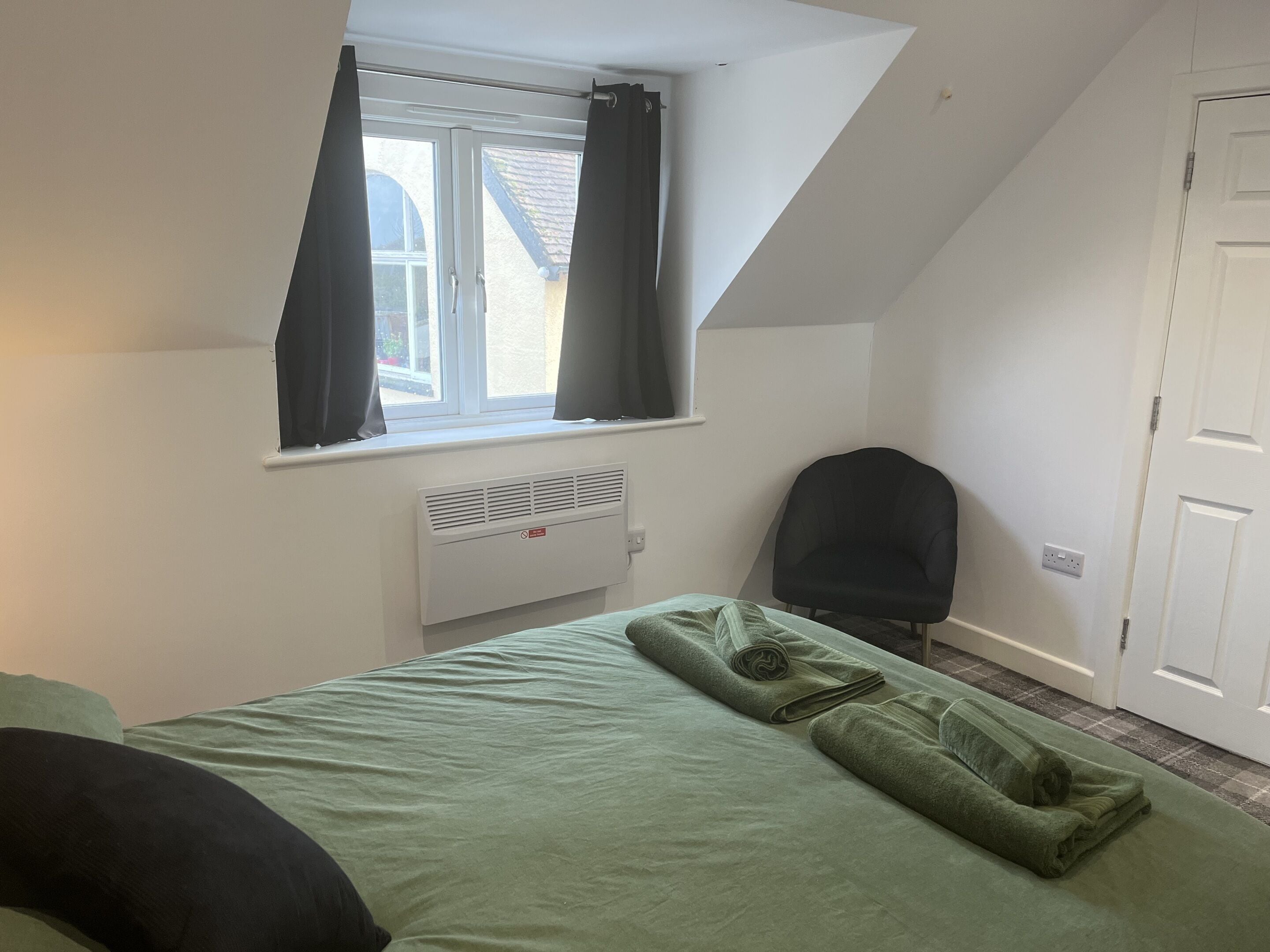A bedroom with a bed, a window with curtains, a radiator, a chair, and a door.