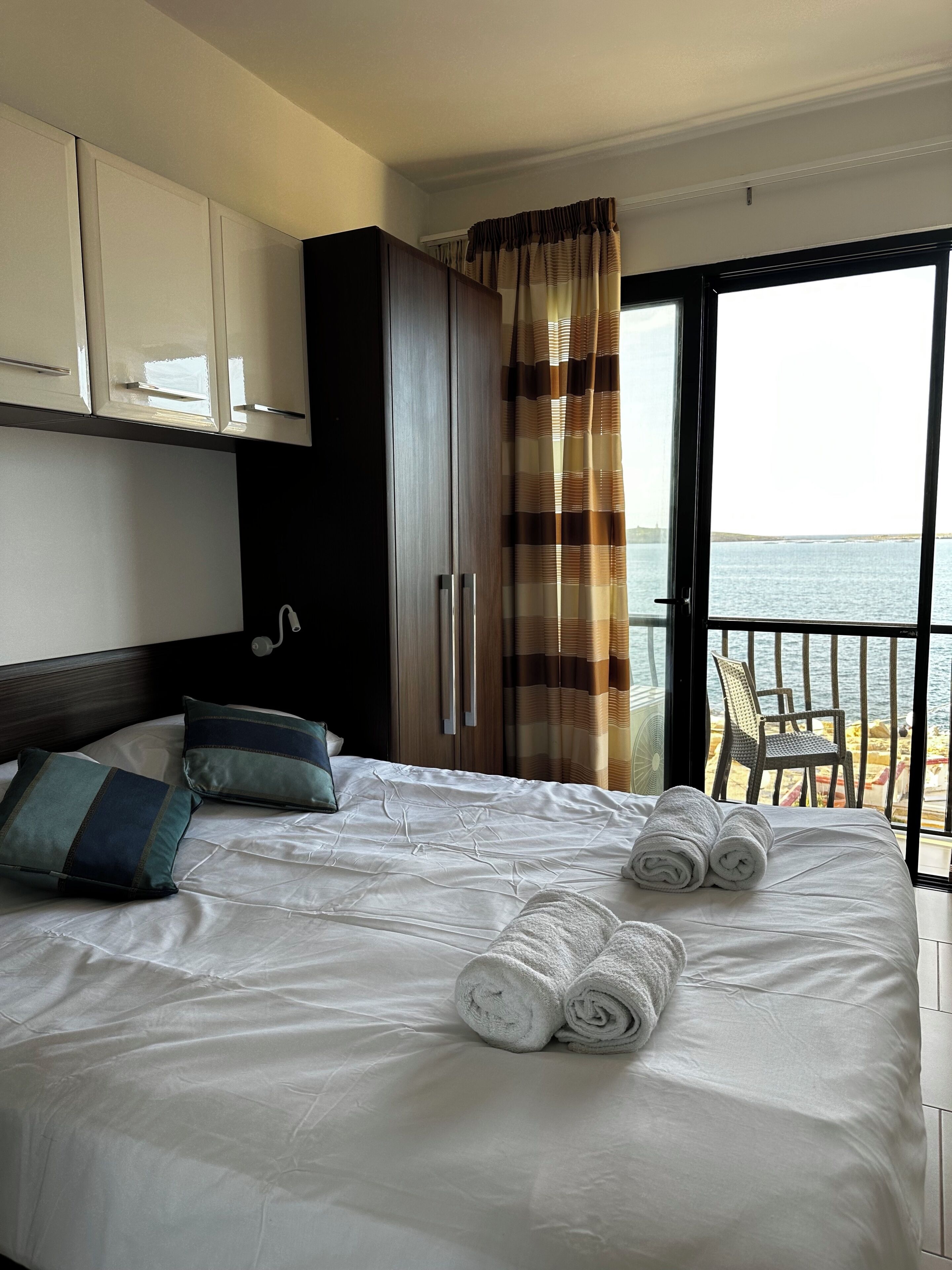 Una camera d'hotel con un letto, cuscini, un armadio, una tenda e vista sul mare.