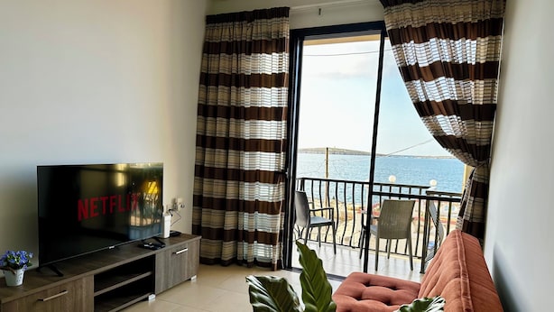 Ett vardagsrum med en TV, en soffa, en balkong med utsikt över havet och gardiner.