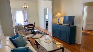A living room with a sofa, a glass coffee table, a television on a cabinet, and a dining area in the background. - Historic Bisbee Retreat - Huge Private Porch -Walk to Town (Bisbee)