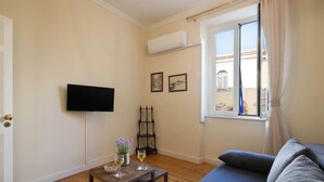 Quarto com uma cama, televisão fixada na parede, uma mesinha com copos e uma planta, e uma janela com cortinas.