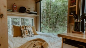 Une chambre confortable avec un lit, des oreillers, une couverture, une fenêtre et une étagère contenant des ustensiles de cuisine.