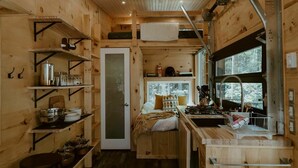 Une petite cabane comprenant un coin cuisine, un lit avec des oreillers et une fenêtre donnant sur les arbres.