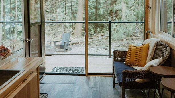 L’intérieur de la cabane est doté d’un plafond en bois, d’un coin repas avec une table et des chaises, d’une cuisine avec un évier et des placards, et d’une grande porte-fenêtre coulissante qui donne sur un espace extérieur boisé.