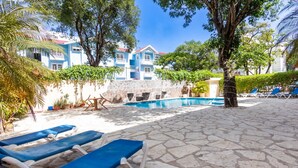 Un área de piscina con sillones de descanso, suelo de piedra y un edificio con fachada azul y blanca.