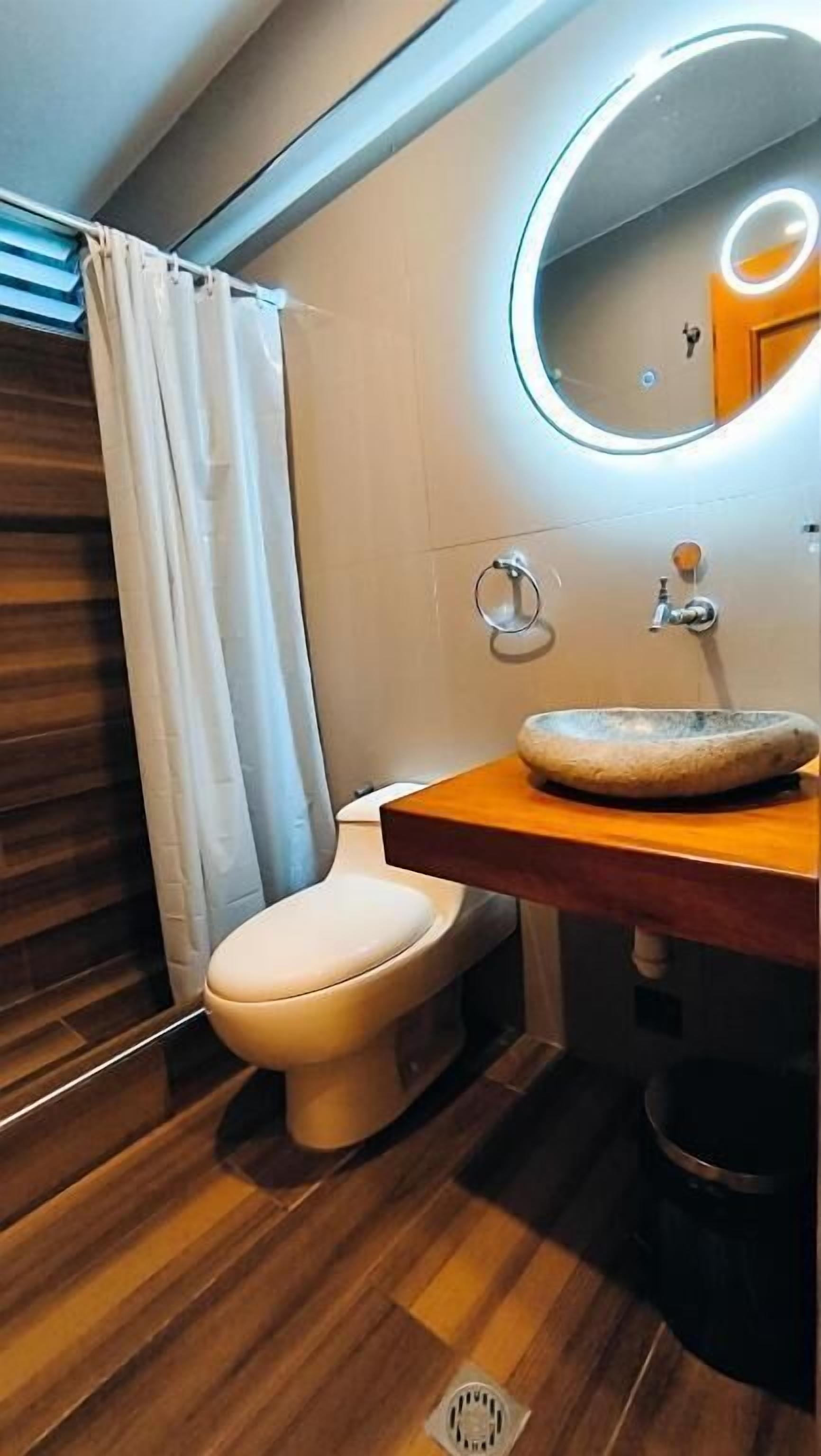 A modern bathroom with a wooden vanity, a round mirror, a toilet, and a shower curtain.