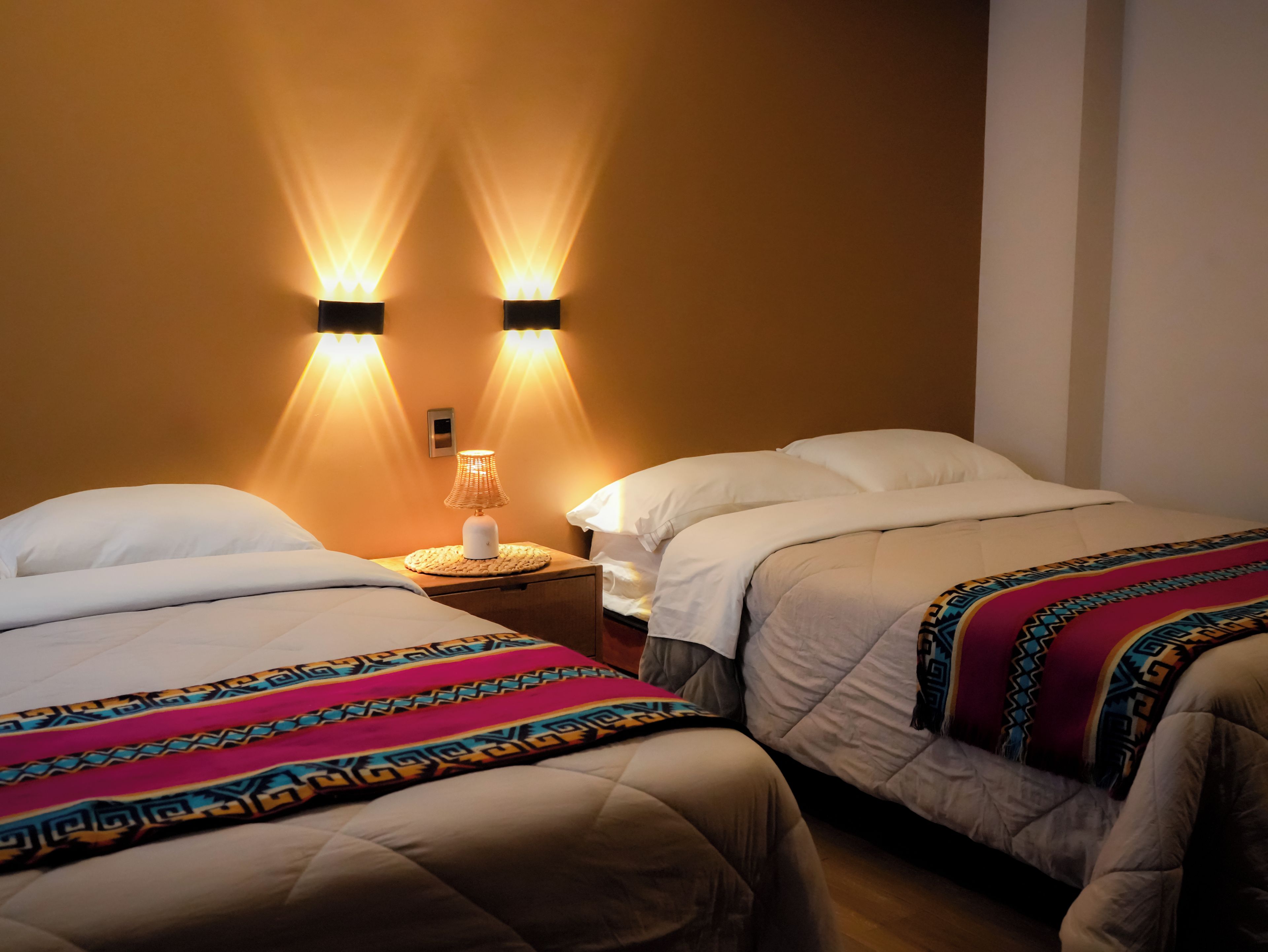 A hotel room with two beds, each with white bedding and a colorful blanket. There is a nightstand with a lamp between the beds.