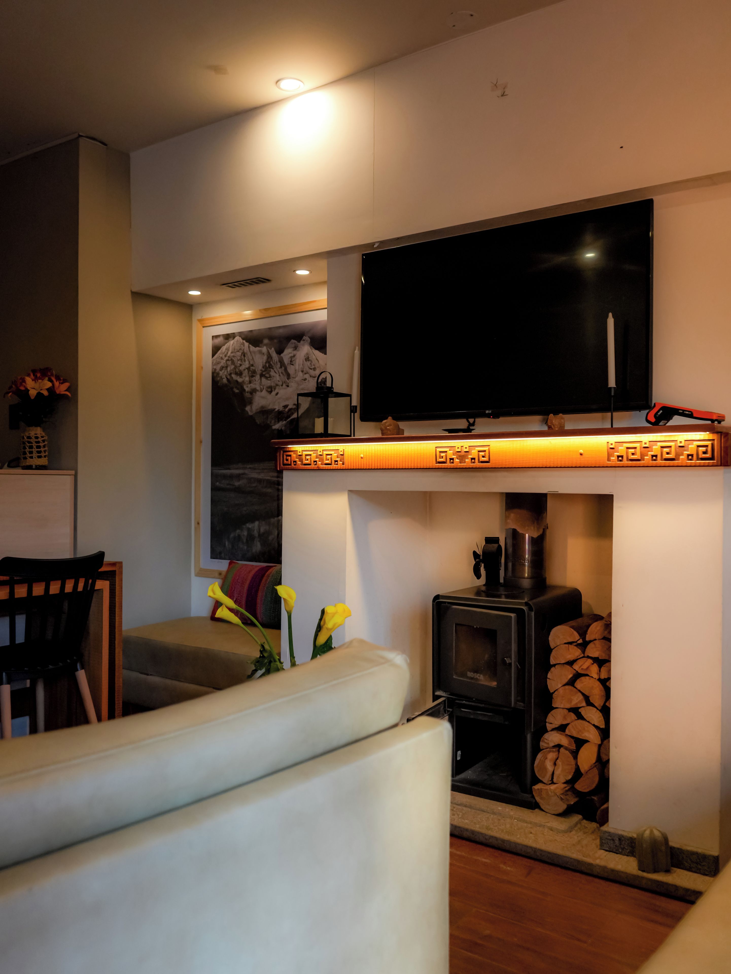 A living room with a sofa, a flat-screen TV mounted above a wood-burning stove, and a decorative shelf with a vase of flowers.