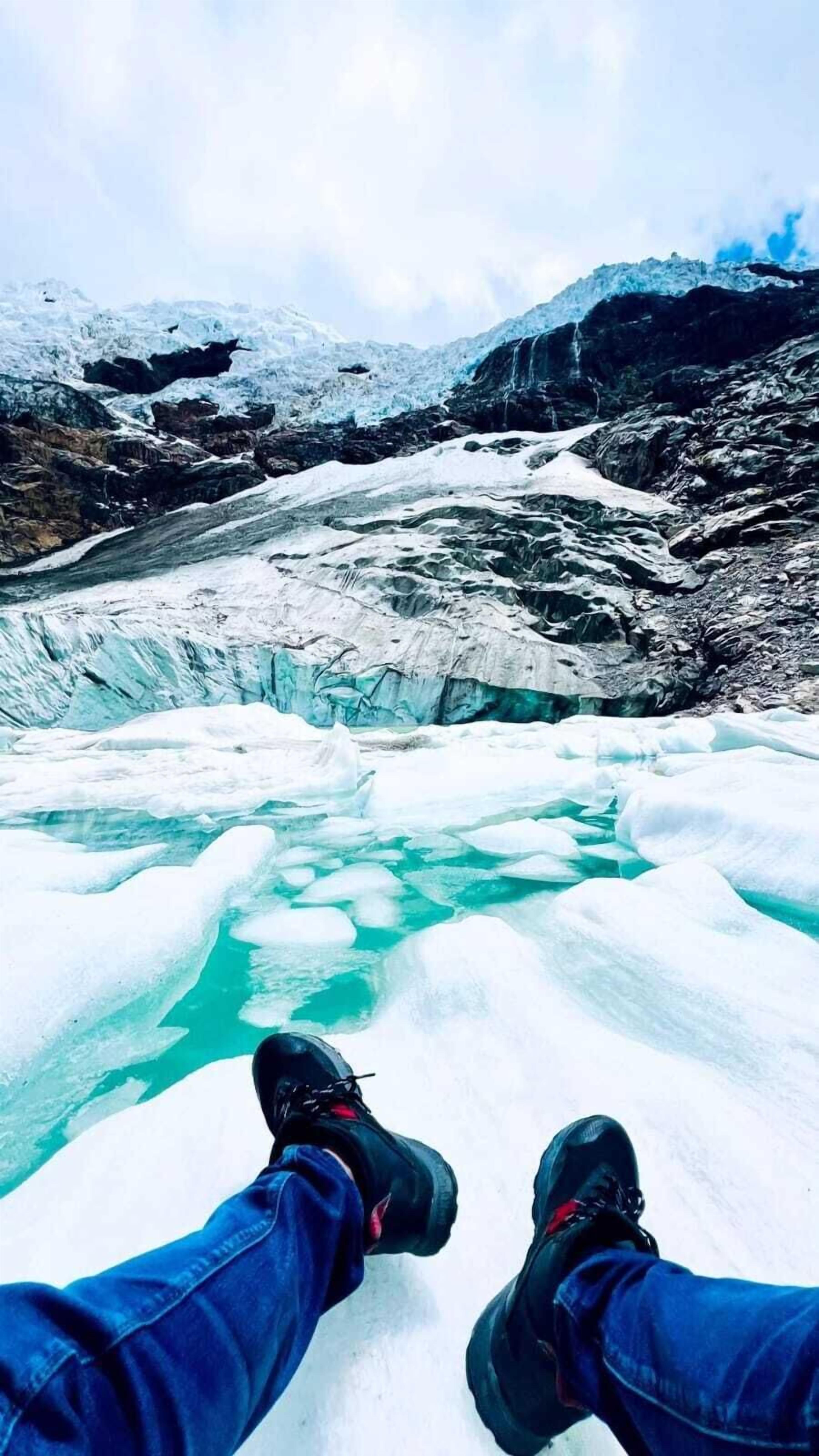A person seated on a snow-covered ledge overlooking a glacier with turquoise water.