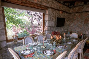 Une table à manger dressée avec des assiettes, des verres et des bougies, entourée de murs en pierre et agrémentée d’une fenêtre donnant sur un paysage verdoyant.