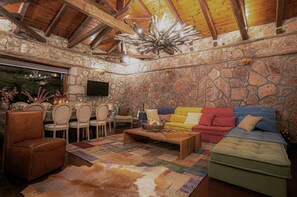 Une pièce aux murs de pierre et au plafond en bois, avec un téléviseur fixé au mur, un coin repas comprenant des chaises et une table, et un canapé aux couleurs vives.