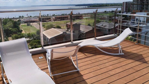 Un balcón con dos sillones blancos, un muelle de madera y vistas a un cuerpo de agua y edificios al fondo.
