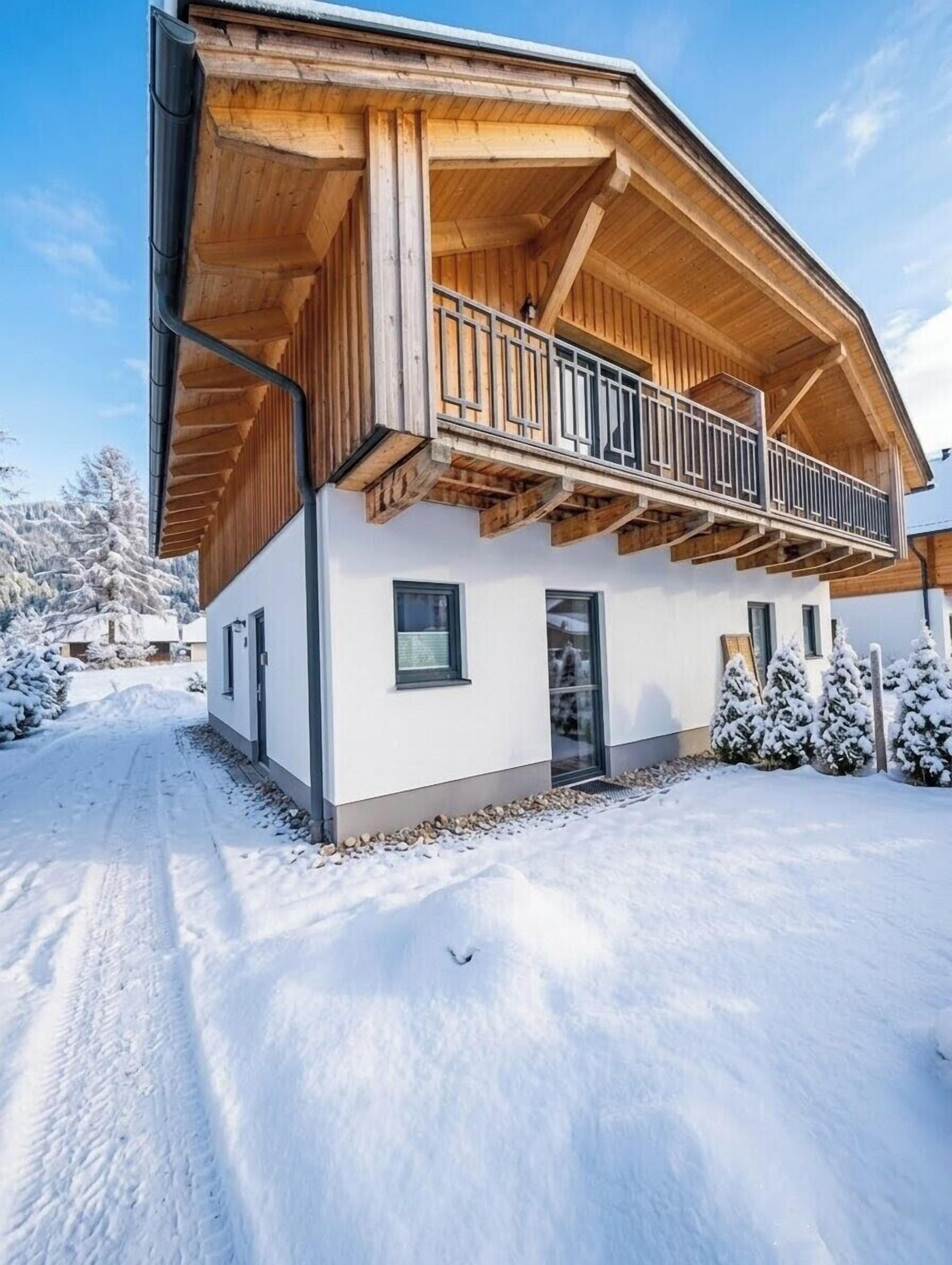 Een modern houten huis met een balkon, een oprit bedekt met sneeuw en omringende bomen.