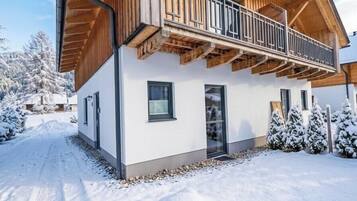 Een modern houten huis met een balkon, een oprit bedekt met sneeuw en omringende bomen.