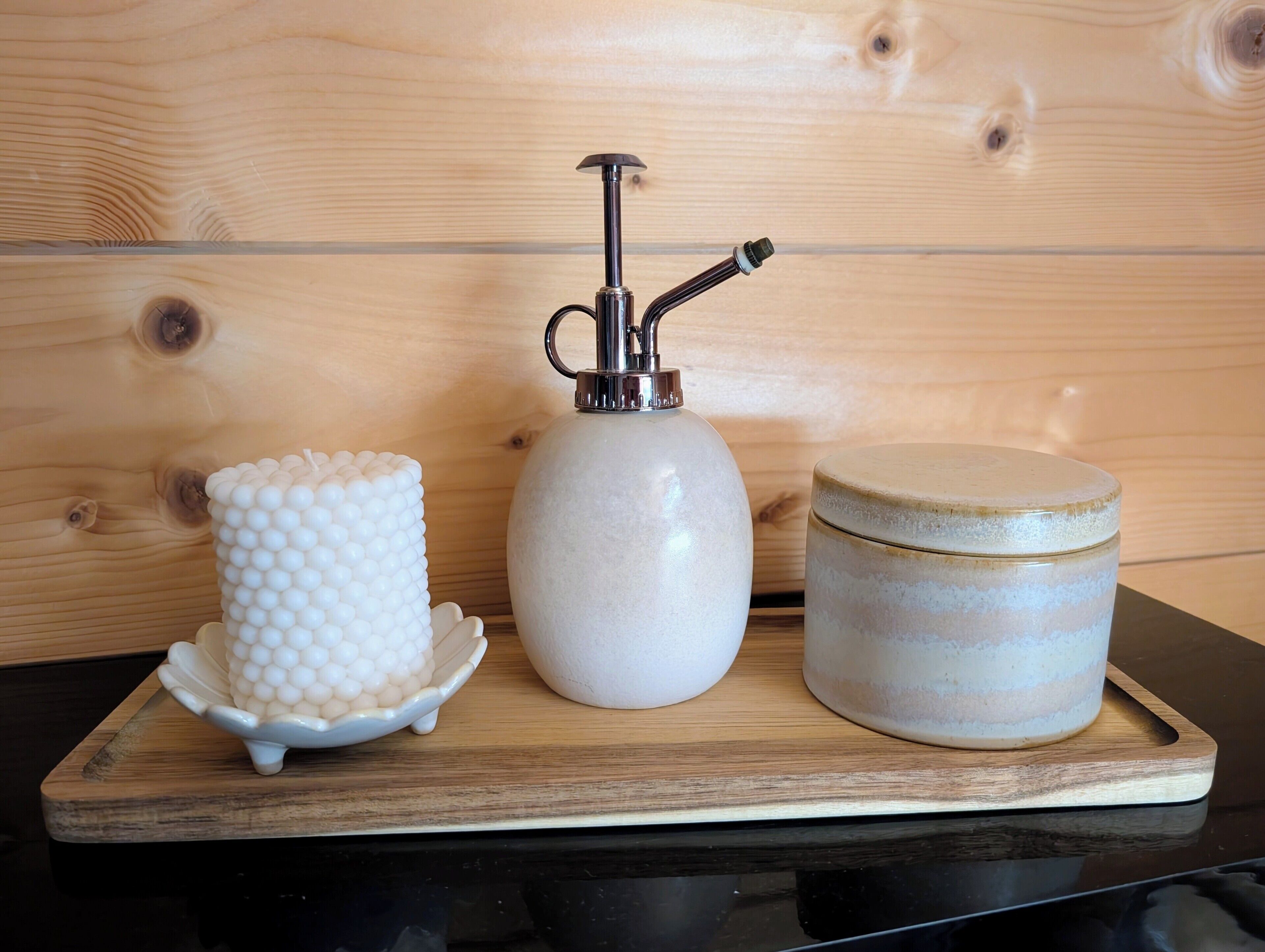 Un plateau en bois contenant trois récipients en céramique : un objet sphérique blanc posé sur une base à motifs festonnés, un objet sphérique blanc muni d’un distributeur, et un récipient cylindrique orné de bandes horizontales.