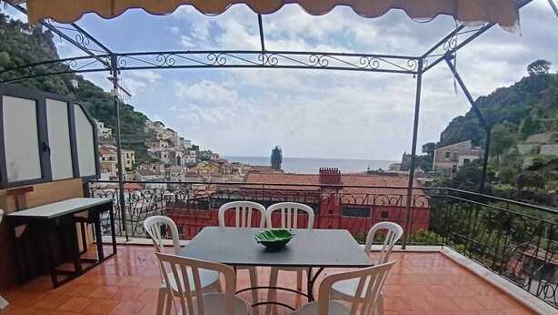 Um terraço com mesa e cadeiras, vista para uma cidade costeira e guarda-corpo de metal.