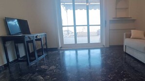 Um quarto com piso de mármore escuro, um sofá bege, uma pequena mesa com um laptop e uma grande janela com vista para uma cerca.