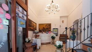 Um hall de hotel com balcão de recepção, um candelabro, uma escadaria e plantas em vasos.