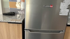 Uma geladeira de aço inoxidável com a marca 'FUJIDENZO' na parte superior, localizada em uma cozinha com armários de madeira e painel de azulejos.