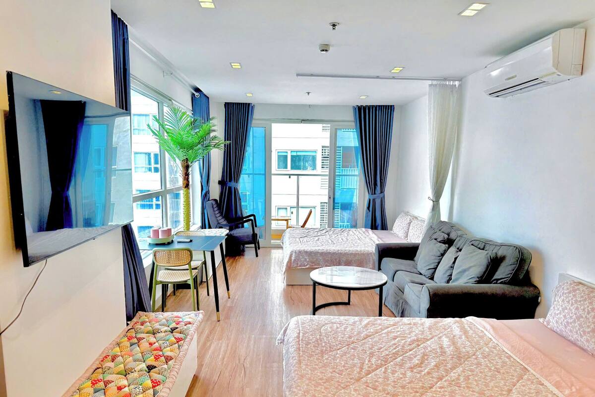 A modern hotel room with a bed, sofa, dining table, and a large window.