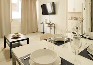 A dining area with a table set for two, a TV mounted on the wall, and a kitchen in the background. - Santa Cruz Holiday Home · 2 Bedrooms & Patio (Santa Cruz de Tenerife)