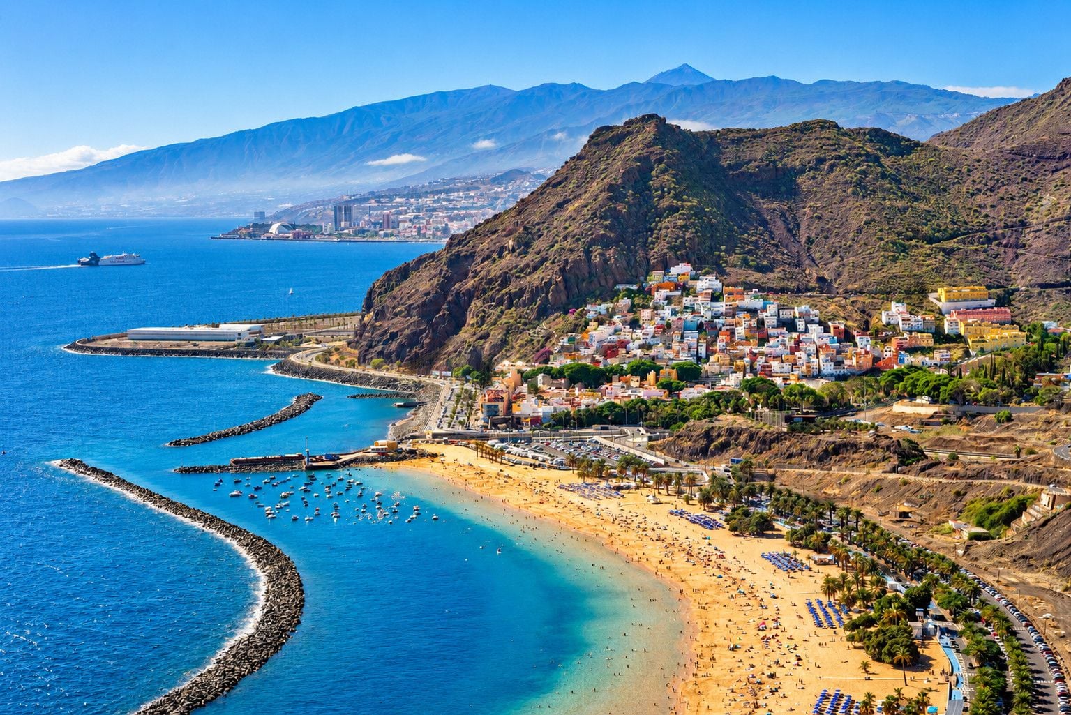 A coastal town with a beach, boats, and a mountainous backdrop.